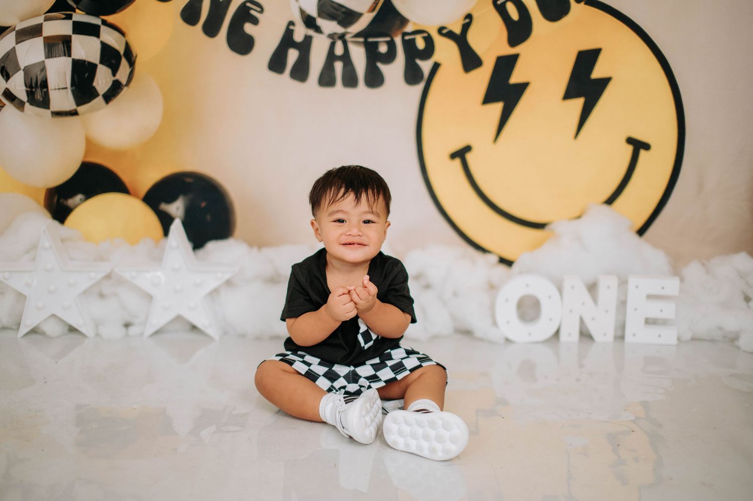 Baby in black and white checkered outfit sits on white floor with yellow smiley face balloon and party decorations.