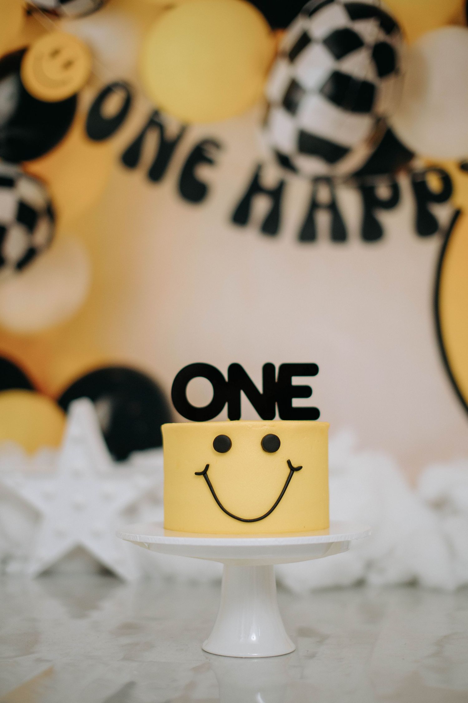 A yellow smiling cake with black 'ONE' topper sits on a white stand with balloon party decorations.