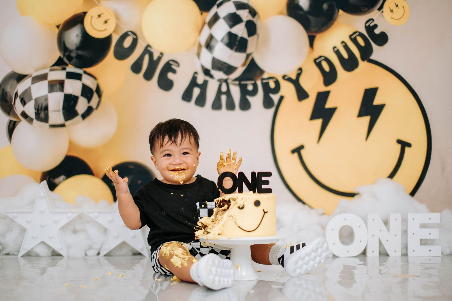 Black and yellow first birthday party setup with checkerboard balloons and smiley face decorations against white background.