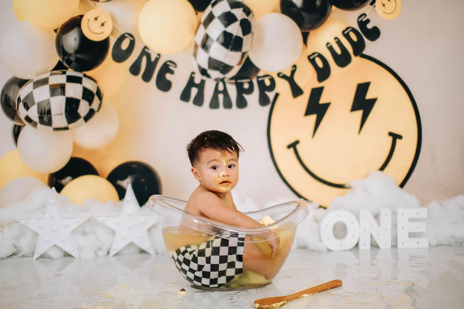 First birthday cake smash photo session with black and white checkered decorations and smiley face balloons against beige backdrop.