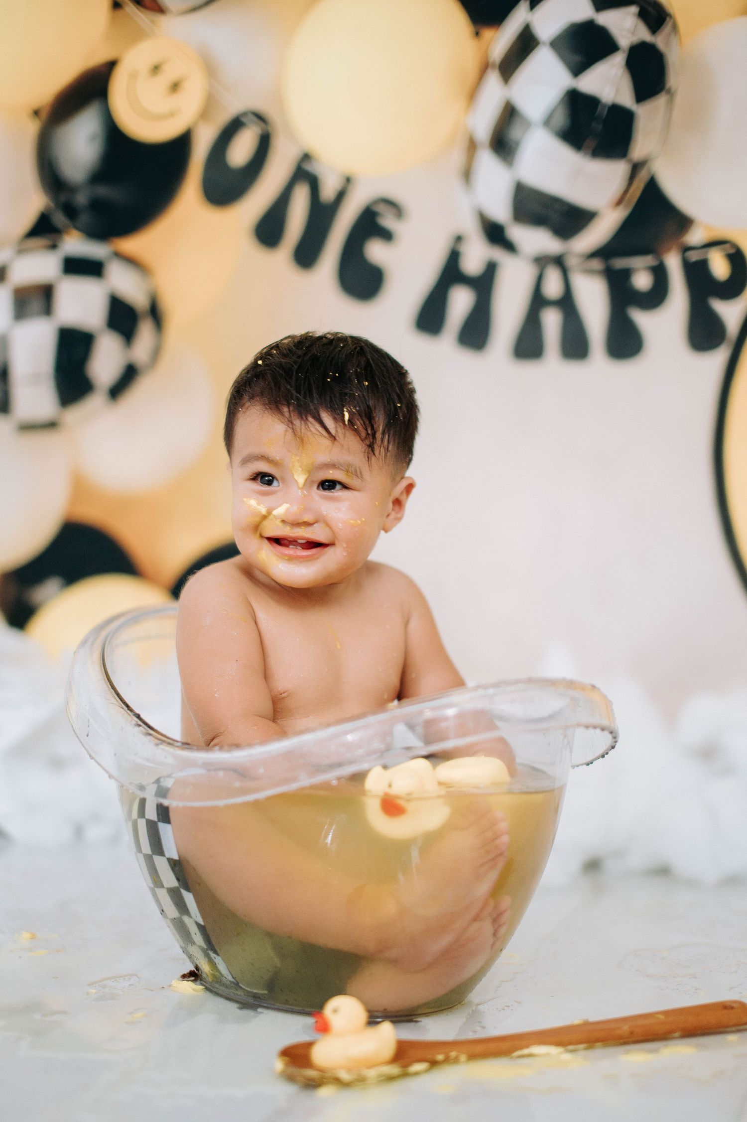 Bath time setup with rubber ducks and clear tub against festive balloon background with 'One Happy' banner.