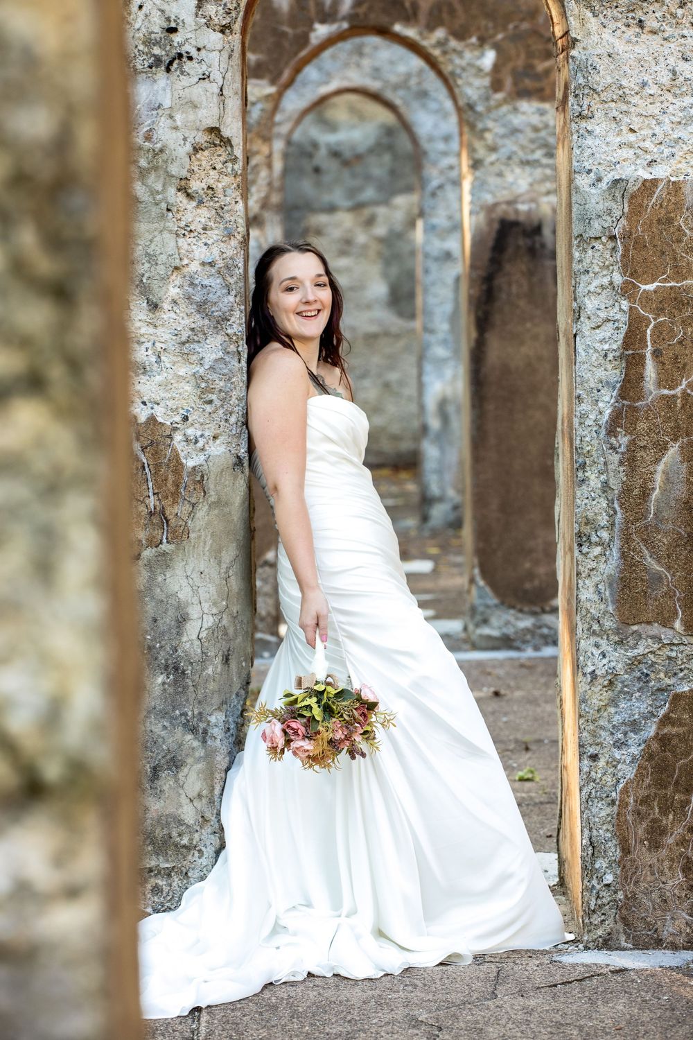 Beautiful bride in white wedding dress holds autumn bouquet while posing in stone archway ruins.