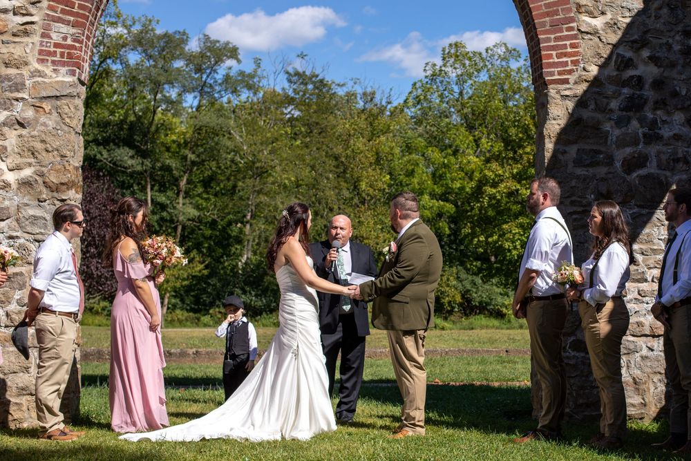 A wedding ceremony takes place between stone archways in a beautiful outdoor garden setting under blue skies.