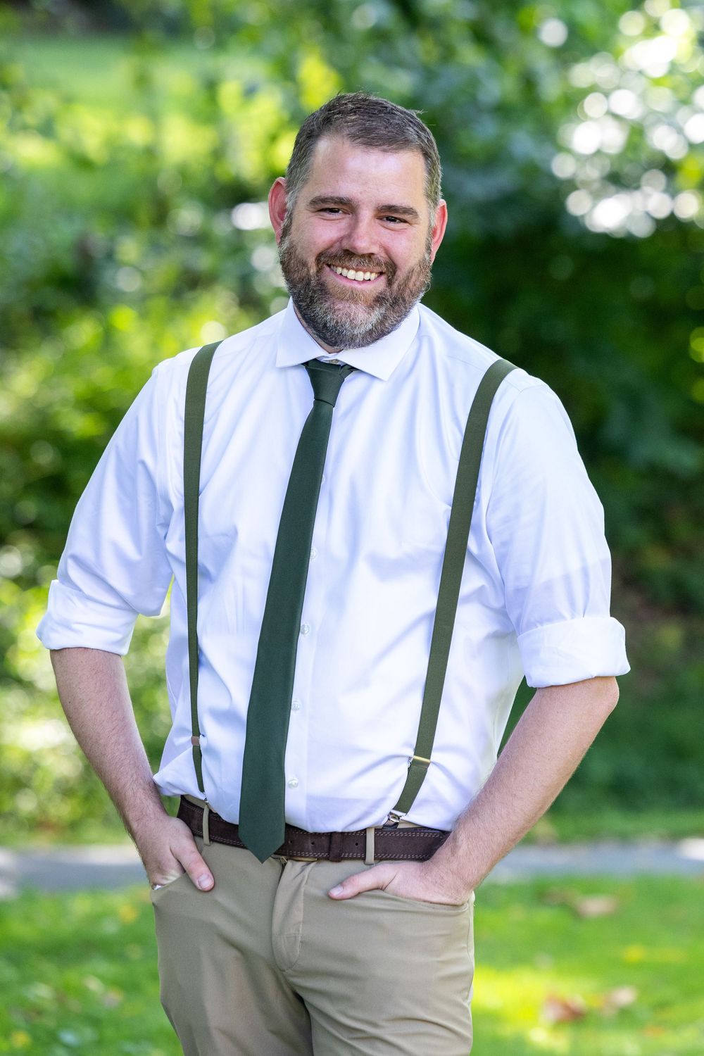 A smiling person in a white dress shirt and green suspenders stands outdoors in a sunny garden setting.