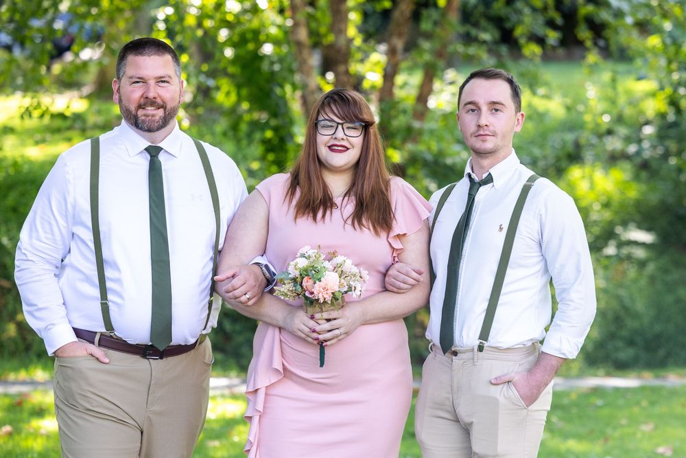 A wedding party in casual attire poses outdoors with suspenders, a pink dress and floral bouquet in a garden setting.
