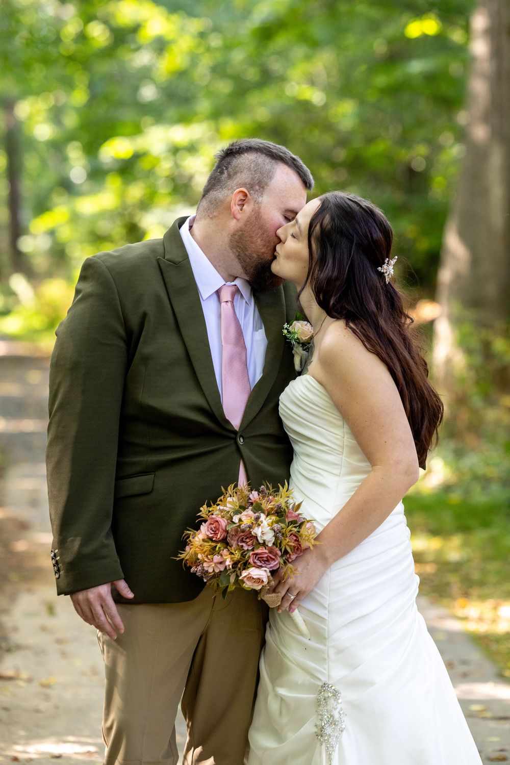 A romantic outdoor wedding photo of a couple kissing in a green forest setting with the bride holding a fall bouquet.