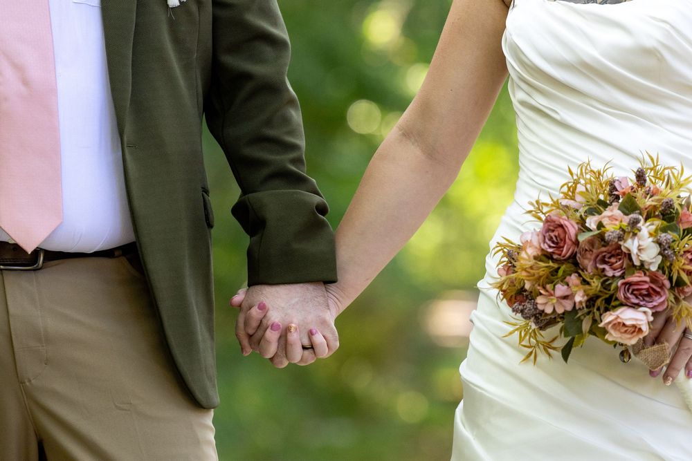 Close-up shot of a couple holding hands at a wedding, with white dress and bouquet of autumn-colored flowers in frame.