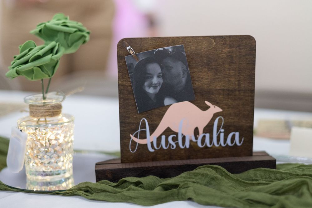 A wooden name display with rose gold lettering sits on a table next to a small mason jar with green leaves and fairy lights.