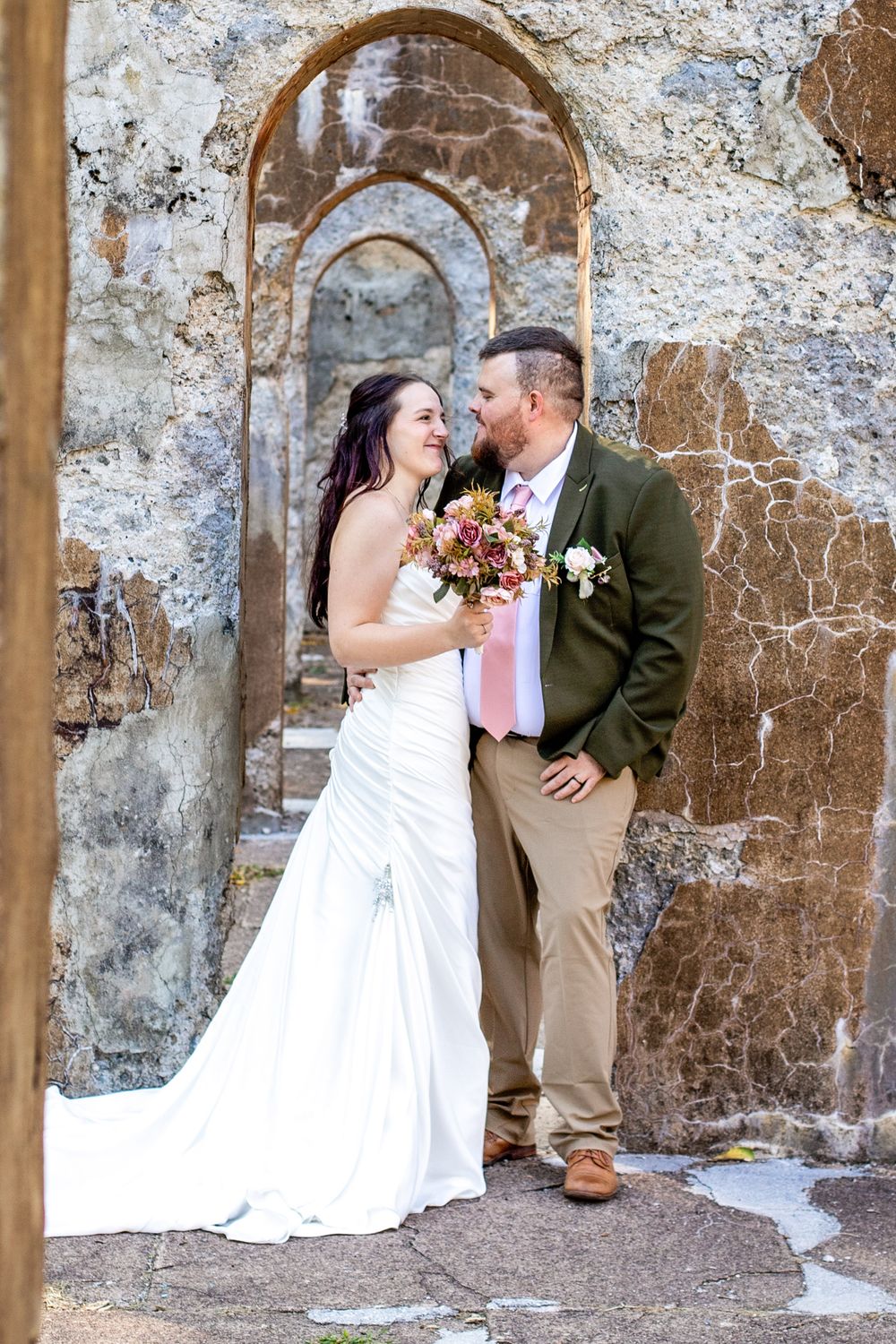 A romantic wedding moment captured in an old stone archway with the couple sharing a tender moment while holding a fall bouquet.
