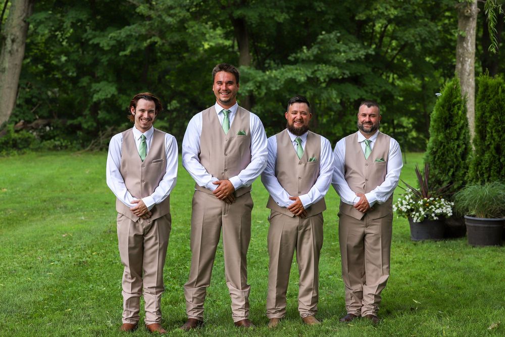 Groomsmen in tan suits stand in a row on lush green grass during an outdoor wedding celebration.