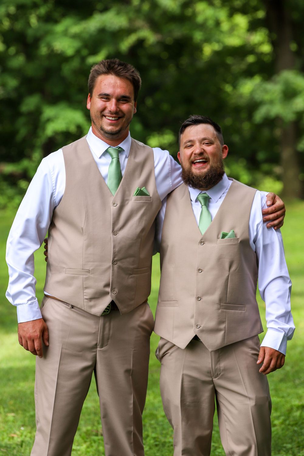 Groomsmen in matching tan vests and mint green ties pose together outdoors against lush greenery.