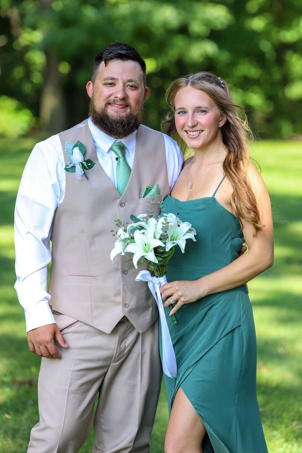 A couple in coordinating green and tan attire pose outdoors holding a white lily bouquet at a formal event.