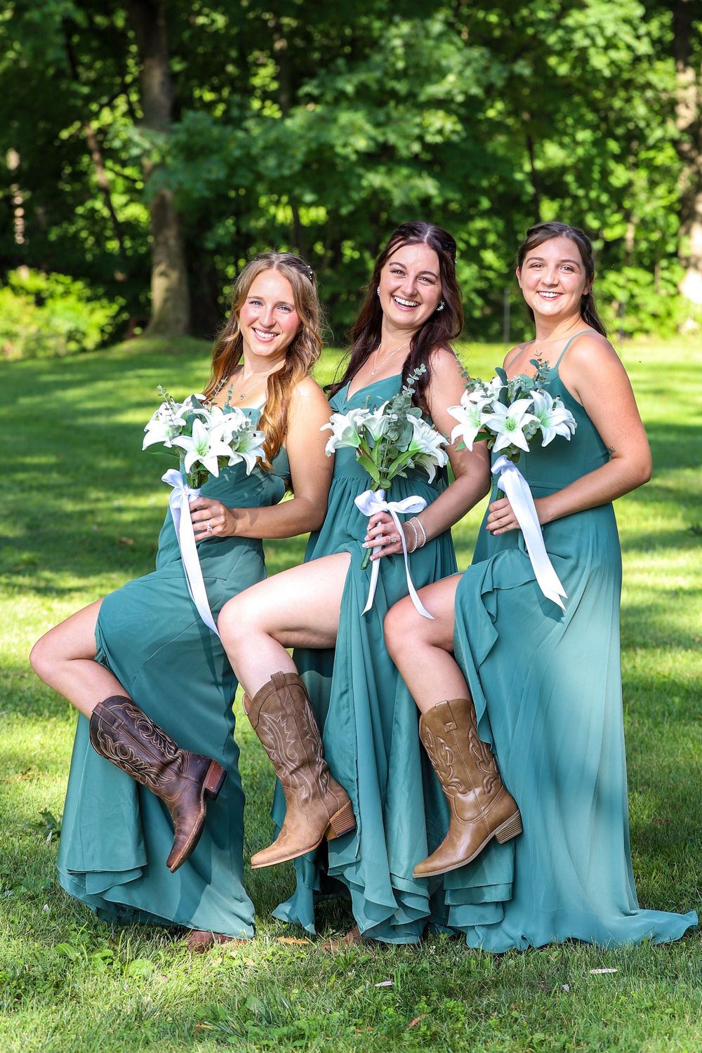 Bridesmaids in teal dresses and brown cowboy boots pose together on grass showing off their western-style footwear.