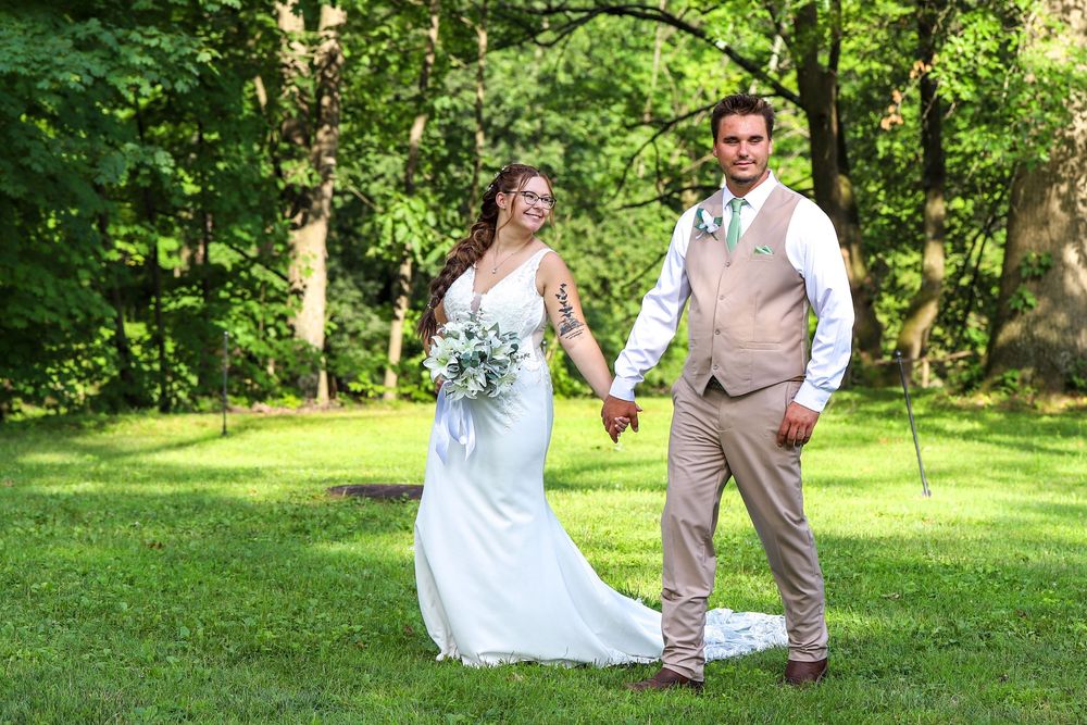A couple in wedding attire walks together through a lush green garden setting on a sunny day.