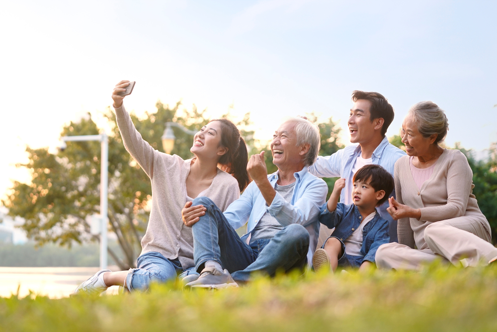 Multi-generational family sitting together on grassy hill during sunset taking a group selfie outdoors.