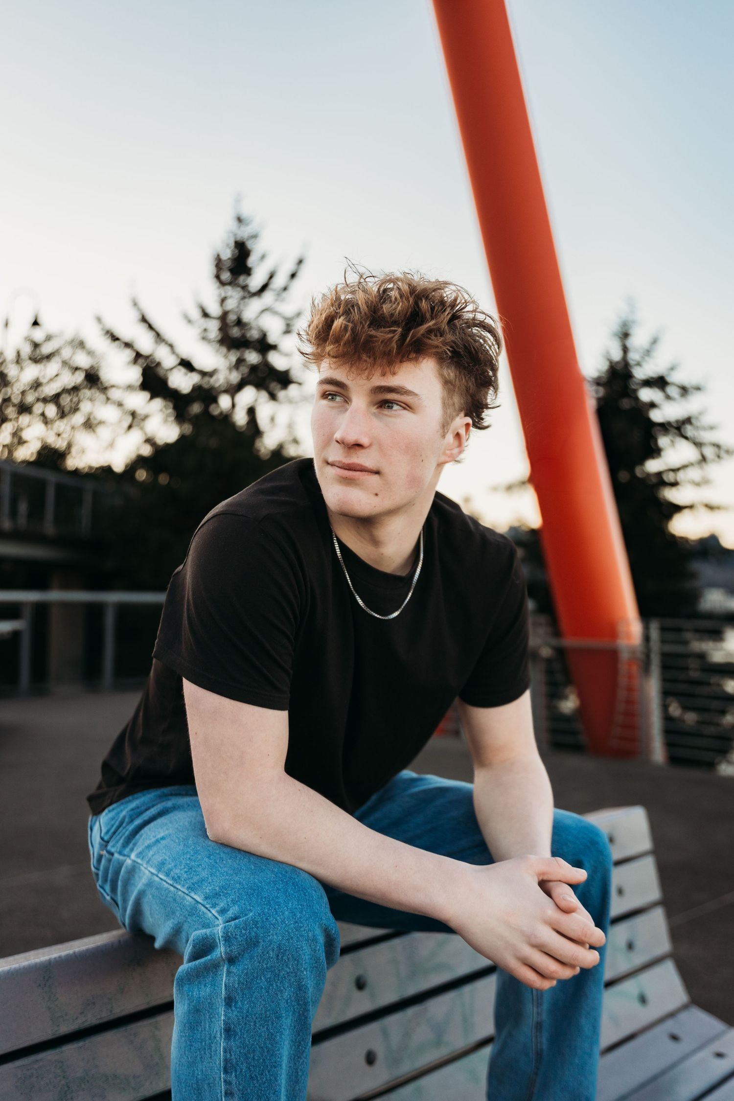 Young person in black t-shirt and jeans sits on bench at sunset with red pole and evergreen trees in background.