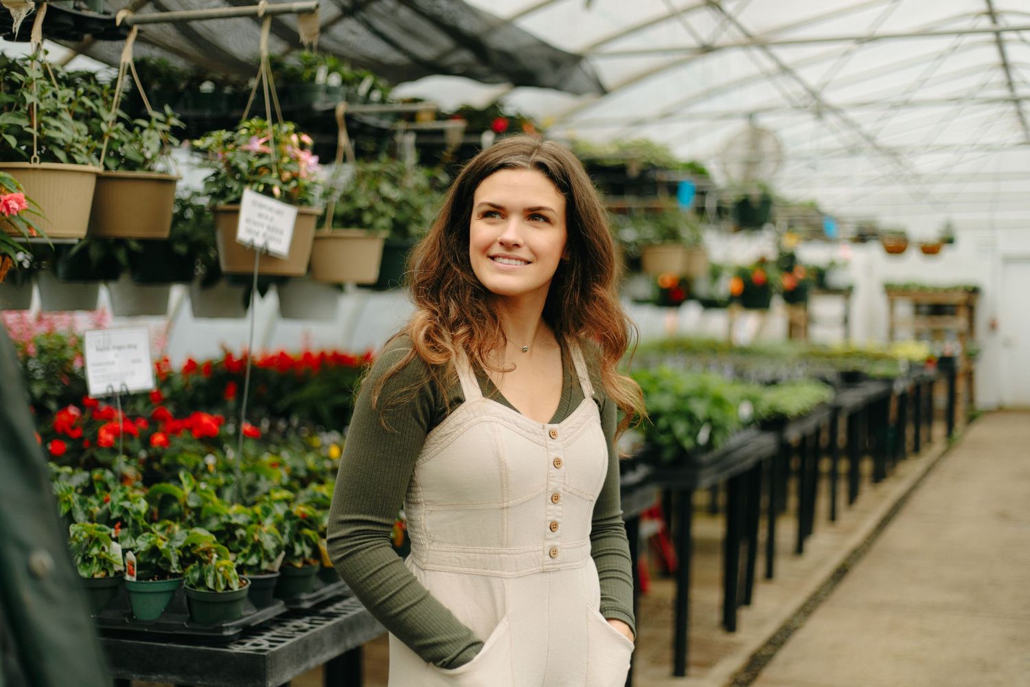 Person in olive cardigan and white dress stands in a greenhouse filled with hanging plants and flowers in pots.
