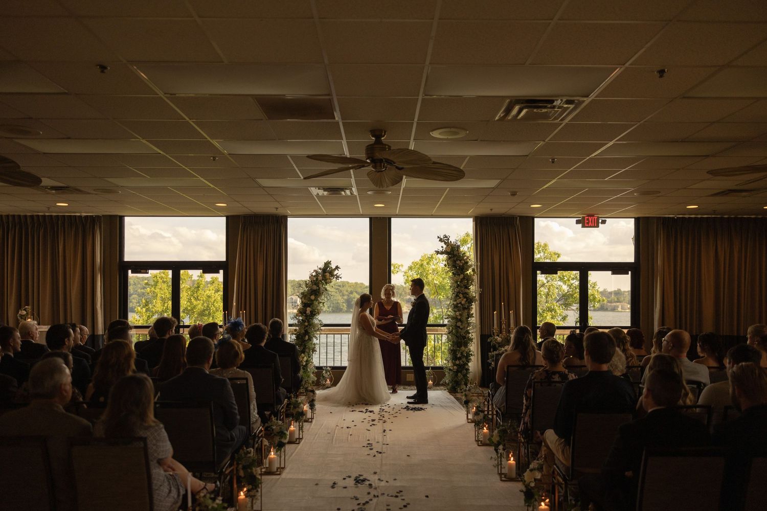 Indoor wedding ceremony with backlit warm sunlight streaming through windows, with guests seated in rows watching the couple.