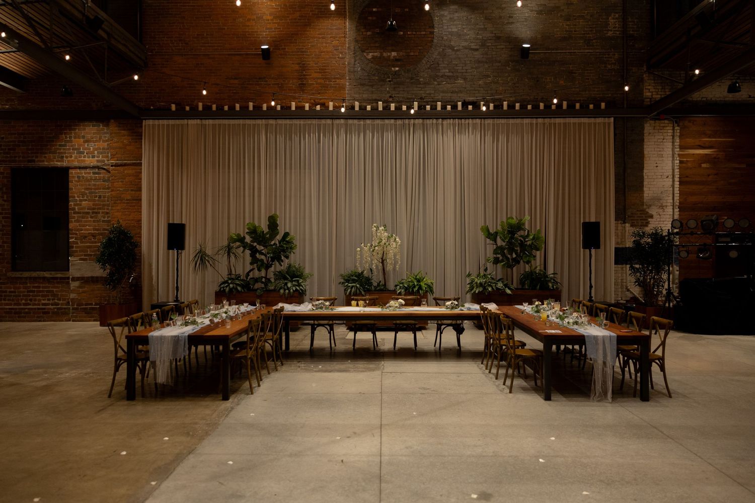 Elegant wedding reception setup with U-shaped table arrangement and white drape backdrop in industrial venue space.