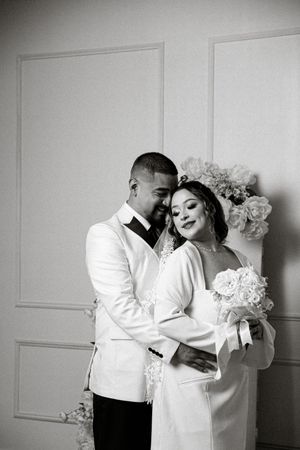 Black and white wedding photo of a couple embracing, bride holding bouquet, groom in white suit, elegant interior backdrop.