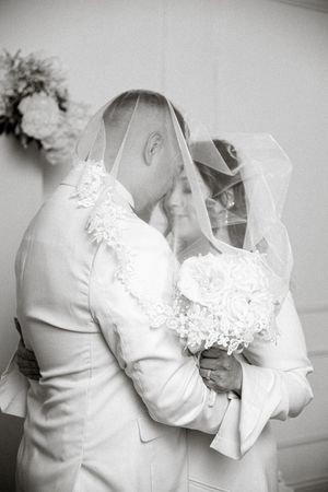 Black and white photo of a bride and groom sharing a kiss under her veil, holding a white bouquet.