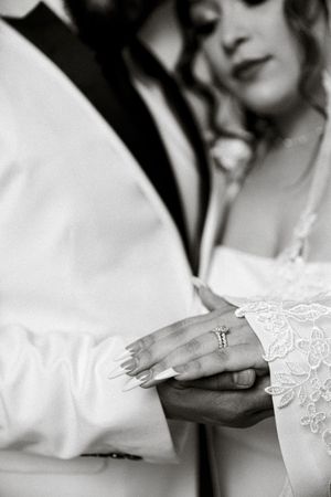 Black and white wedding photo of bride and groom embracing, showcasing her diamond ring and lace dress sleeve.