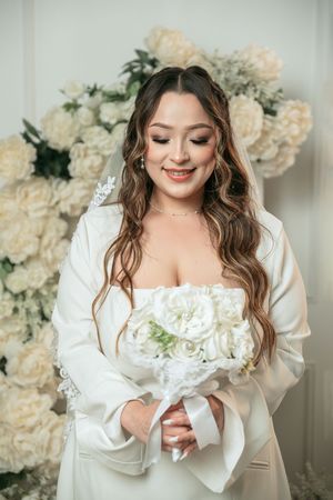Smiling bride in white wedding dress holding bouquet of white roses, standing before floral backdrop.