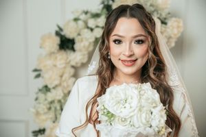 Smiling bride in white dress and veil holding bouquet of white flowers with floral backdrop.