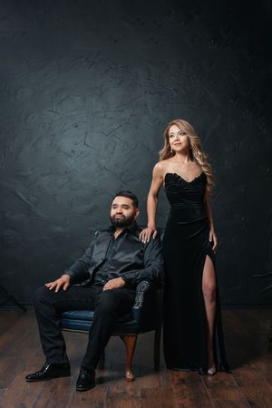 Elegant couple in black formal attire posing in studio, man seated in chair, woman standing beside him in velvet gown.