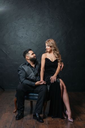 Elegant couple in black formal attire posing in a dark studio, woman in velvet gown sitting beside suited man.