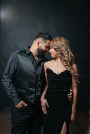 Elegant couple in matching black outfits sharing a romantic moment against a dark textured wall.