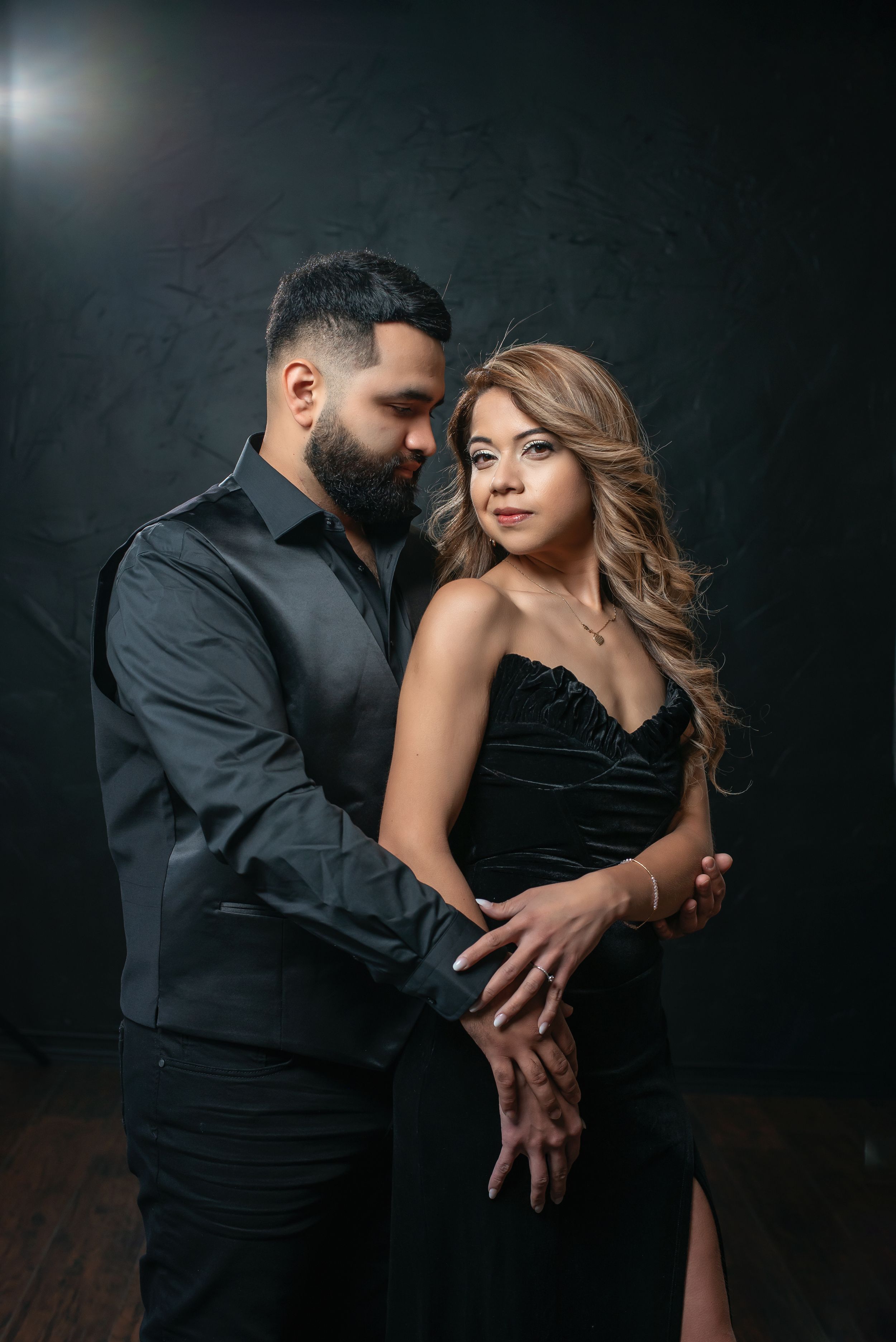 Elegant couple in matching black outfits embracing in a dark studio, woman in strapless gown, man in black shirt.