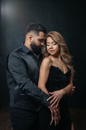 A couple in elegant black outfits embrace intimately in a dramatic dark studio setting.