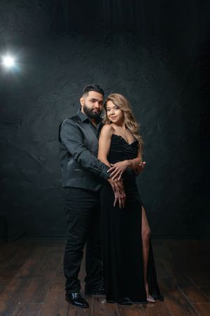 Elegant couple in matching black outfits posing together in a dark studio, woman in a slit gown, man in a suit.