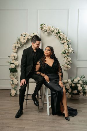Elegant couple in matching black formal attire posing before a white floral arch with roses in a bright studio setting.