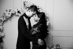 Black and white photo of a romantic couple in elegant attire embracing before a floral arch backdrop.