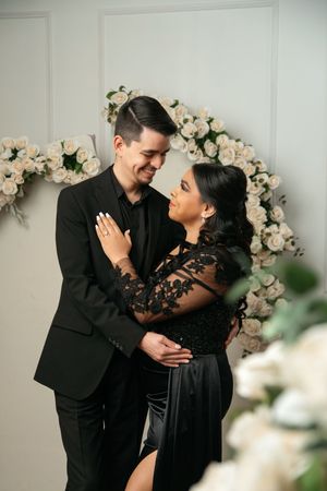 Couple in matching black formal attire embracing before a white floral arch backdrop in an elegant engagement photo.
