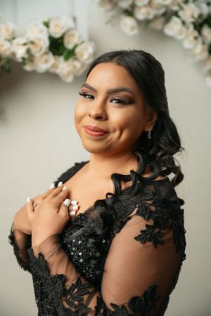 Woman in black lace dress with elegant makeup poses gracefully in front of white rose floral backdrop.