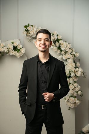 Young man in black suit smiling, standing before white rose floral arch backdrop.