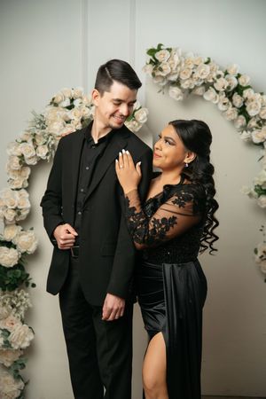 Elegant couple in matching black formal attire posing before a white floral arch, woman placing hand on mans chest.