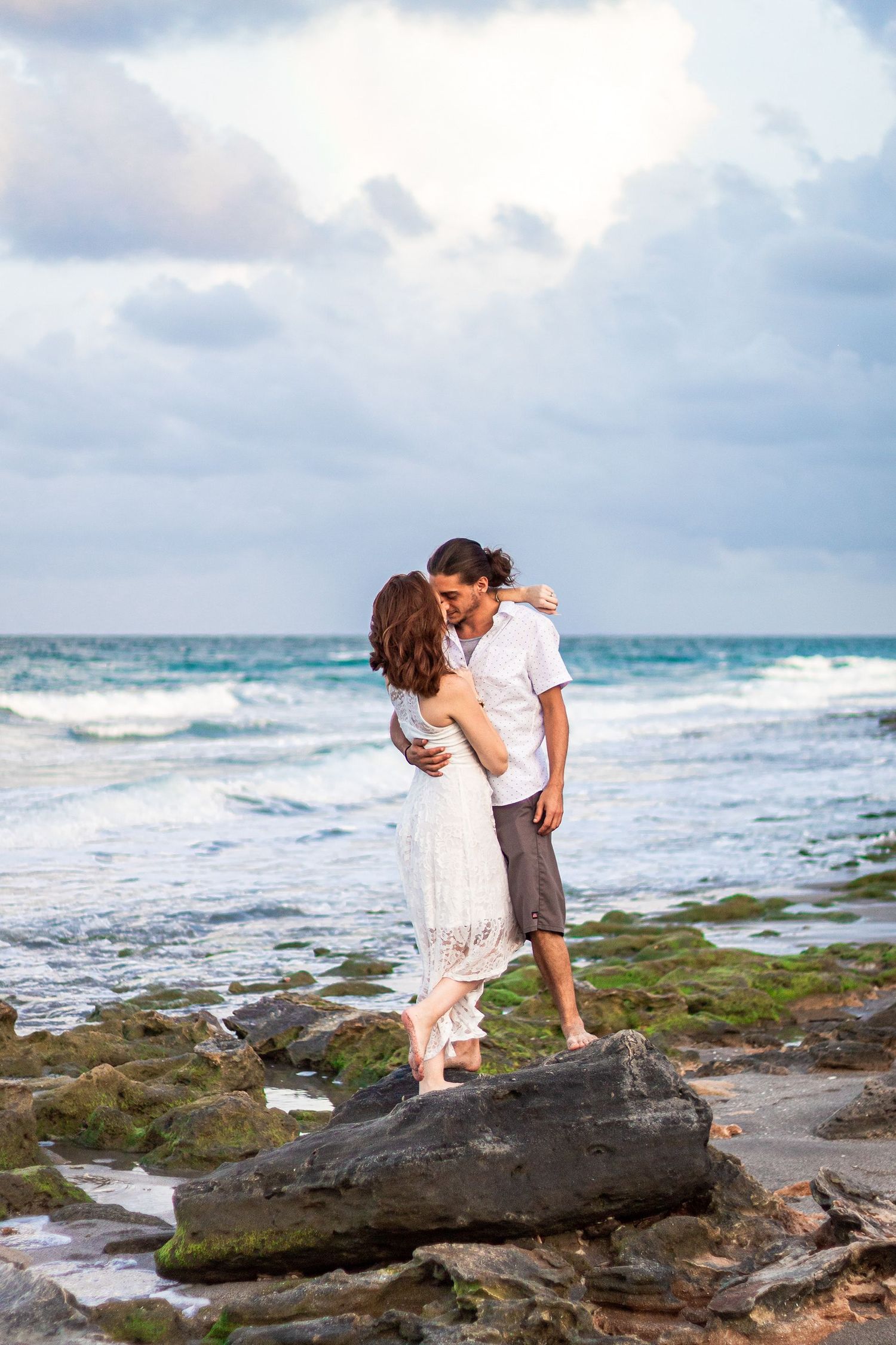 A romantic couple embracing on rocky beach with waves crashing in the background.