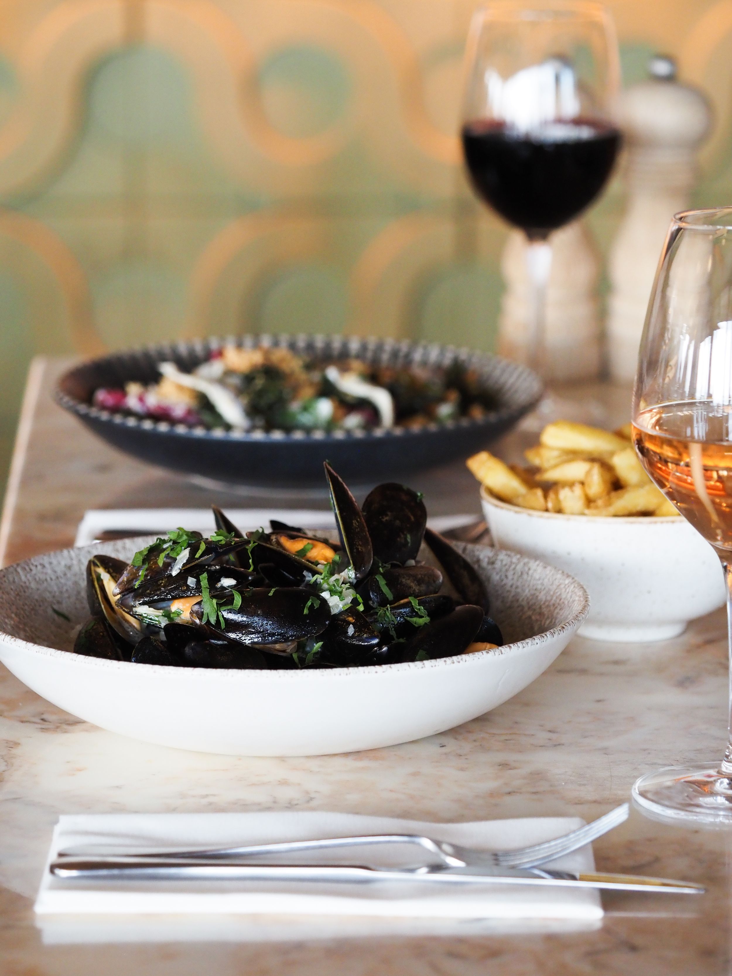Steamed mussels in a white bowl served with crispy fries and red wine at an upscale restaurant table.