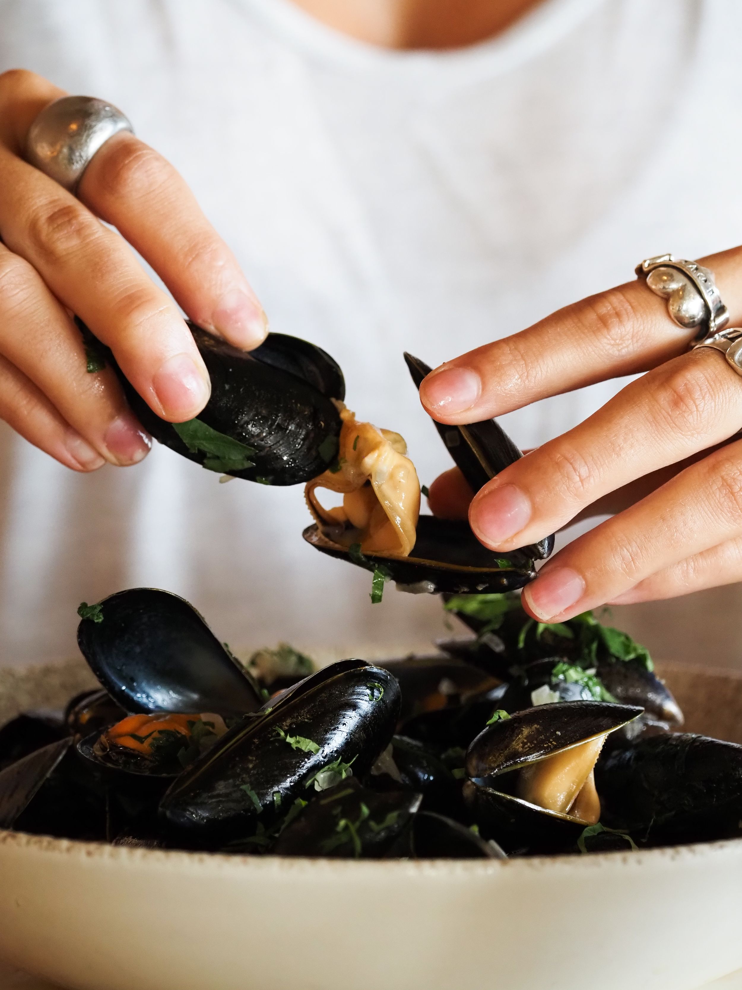 Hands using mussel shell to scoop fresh steamed mussels from a white bowl, wearing silver rings.