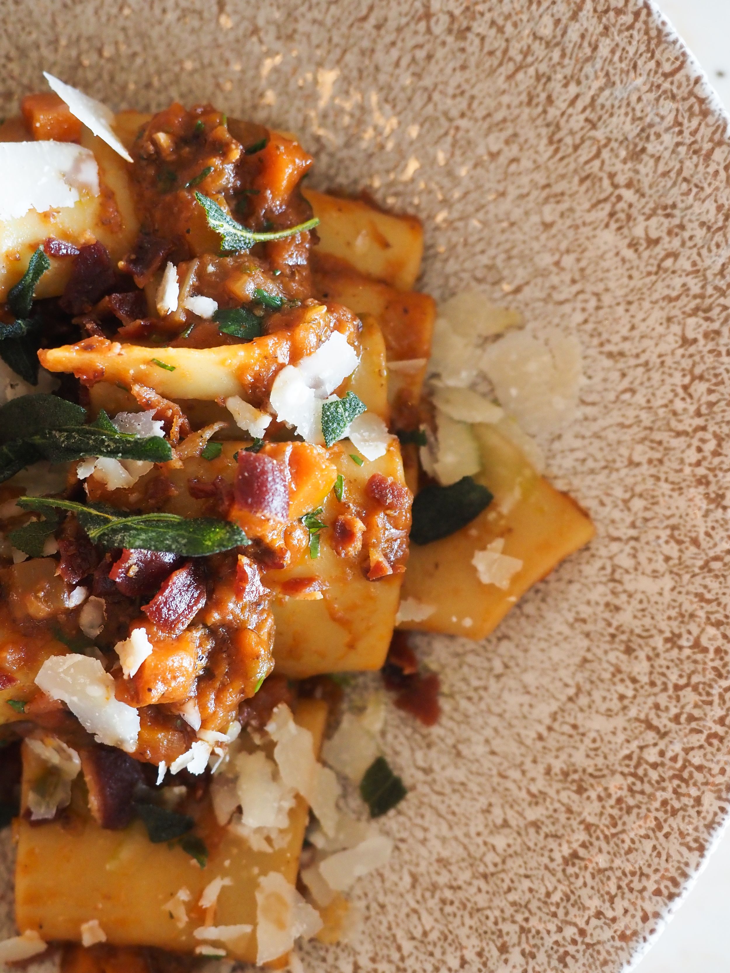 Close-up of pasta dish with tomato sauce, crispy bacon, fresh herbs, and shaved Parmesan in a rustic bowl.