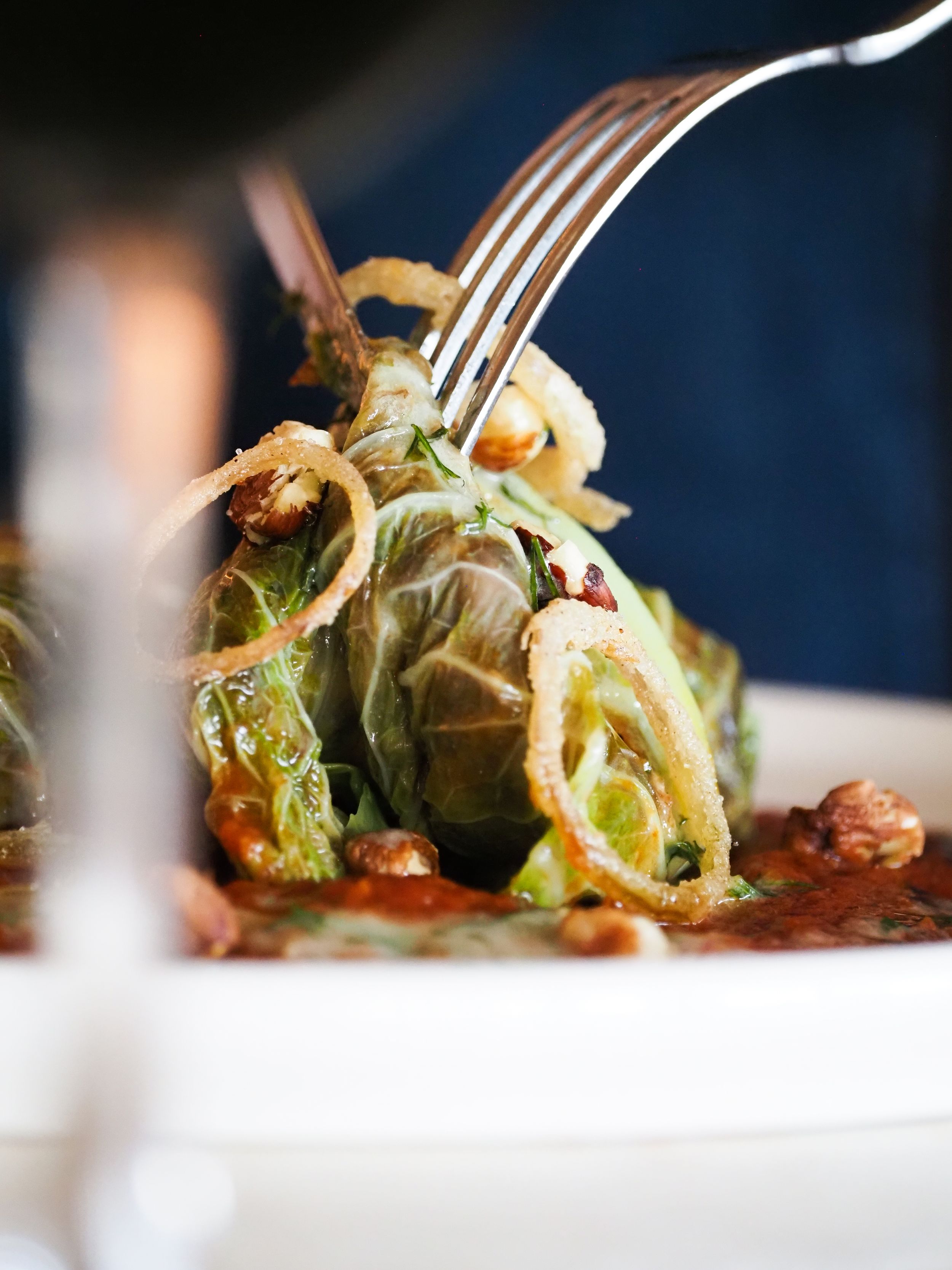 Fork piercing a stuffed cabbage roll in tomato sauce, topped with crispy onions on a white plate.