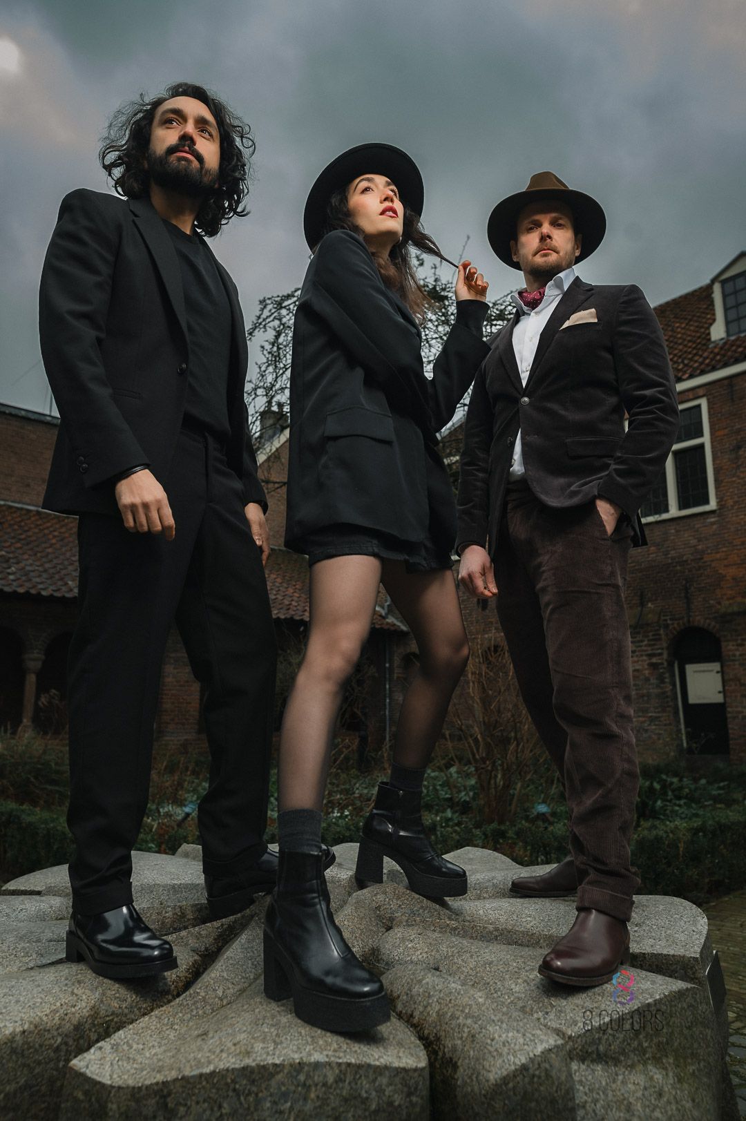 Low-angle shot of three musicians in dark attire posing on concrete steps with dramatic cloudy sky backdrop.