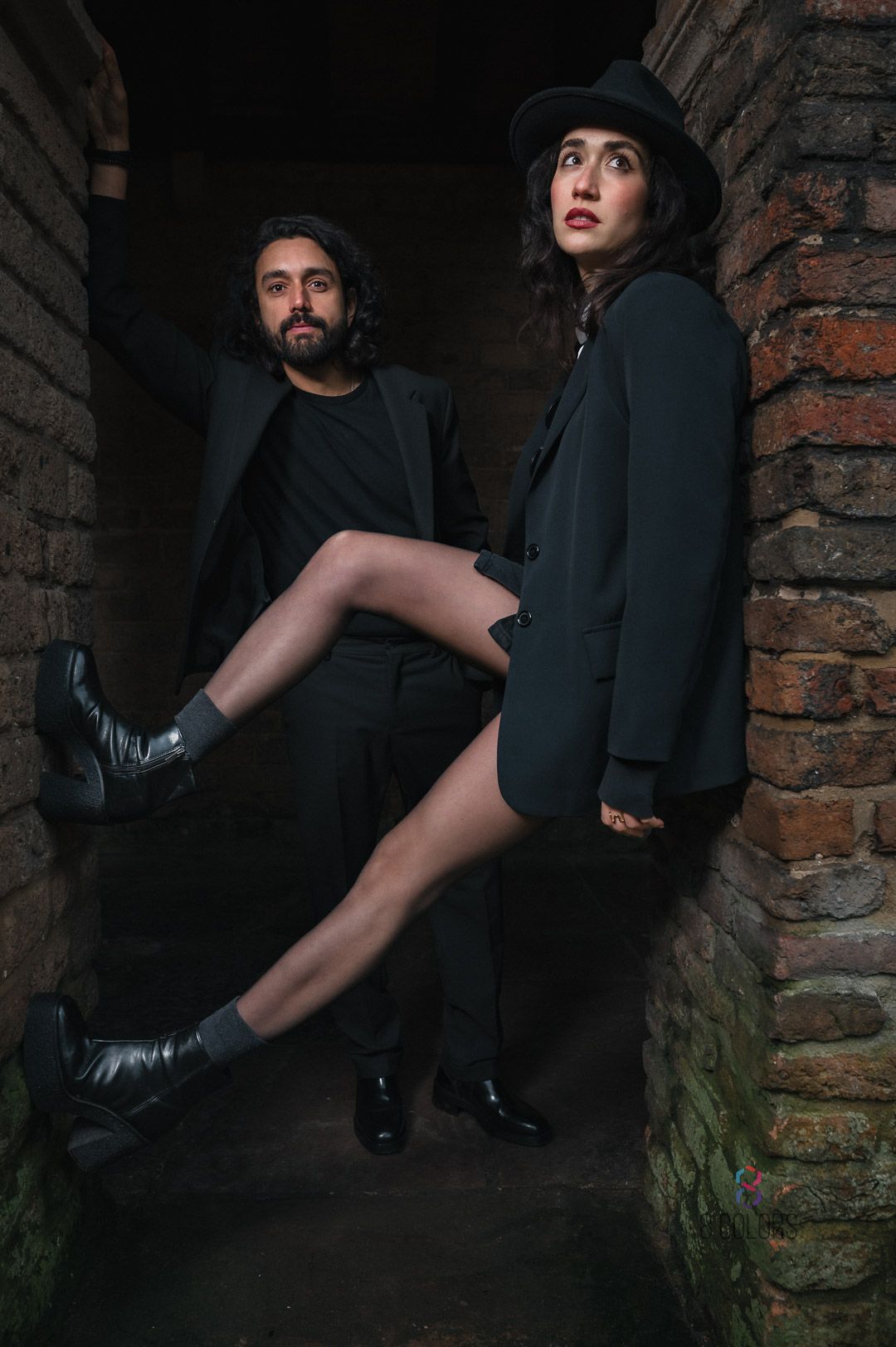 Artistic fashion portrait of two people in dark clothing and boots posing dramatically against brick wall in moody lighting.