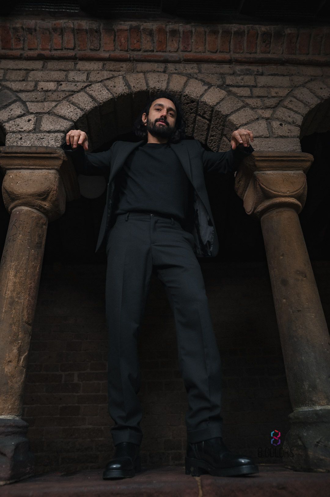 Low angle shot of a person in dark formal wear posing between stone columns of a gothic architectural entrance.