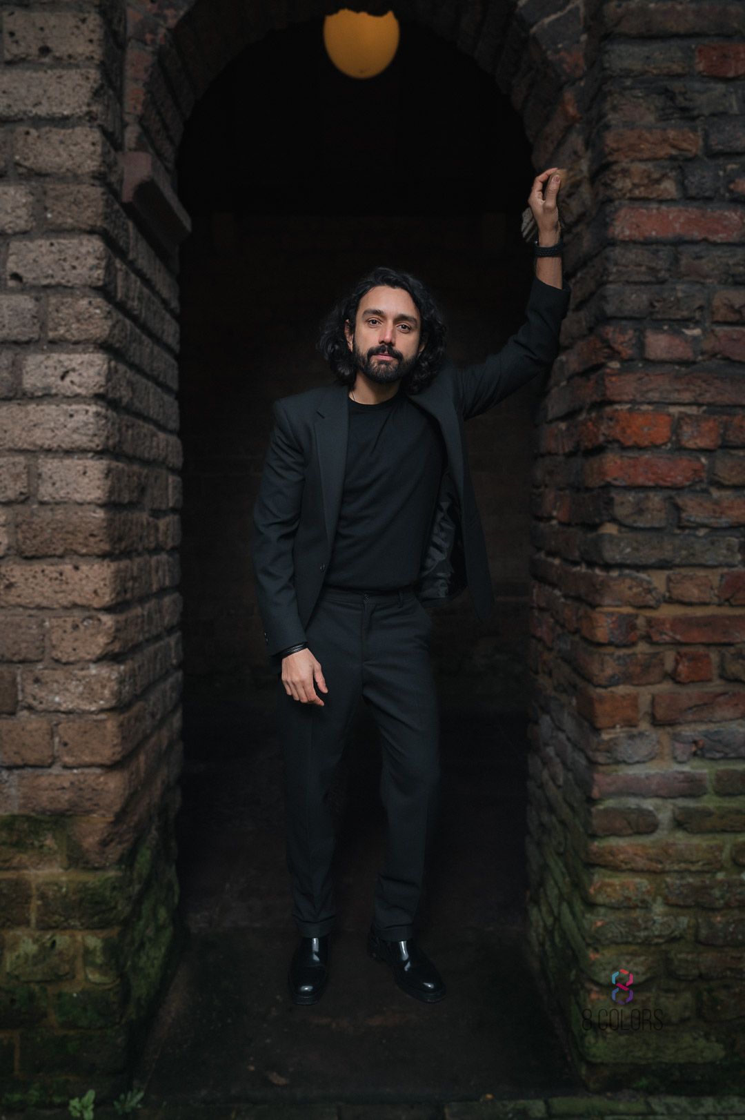 A person in a dark suit poses within a moody brick archway beneath an orange light.