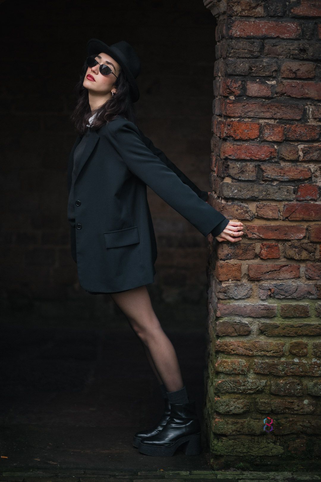 Dramatic fashion photo of figure in black blazer and platform boots leaning against brick wall in moody lighting.